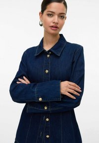 Femme aux cheveux plaqués en arrière portant une veste en denim bleu foncé avec des boutons dorés et des boucles d'oreilles en argent, les bras croisés sur la poitrine.