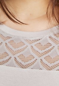 Ljusgrå topp med ett texturerat hjärt-mönster på ett genomskinligt tyg. Slät halsringning med en kontrasterande enfärgad kant.