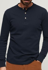 Marineblå langærmet henley-skjorte i bomuld med flad krave, tre-knap placket og sømdetaljer i sidesømmen.