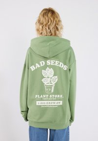 Grön hoodie med en stor vit grafik med texten "BAD SEEDS" och en växtillustration på baksidan. Mjuk tyg, avslappnad passform.