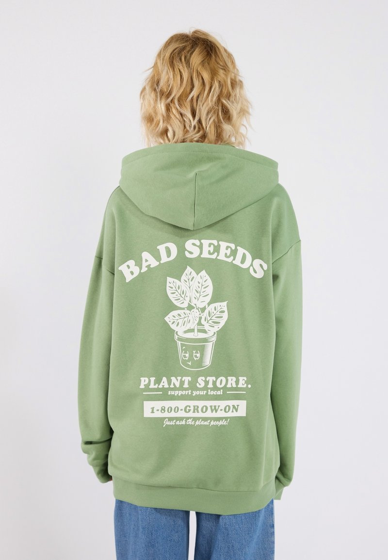 Grön hoodie med en stor vit grafik med texten "BAD SEEDS" och en växtillustration på baksidan. Mjuk tyg, avslappnad passform.
