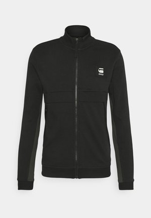 Veste noire zippée intégrale avec col, poignets et bas côtelés, poches latérales et petit patch logo blanc sur la poitrine gauche.