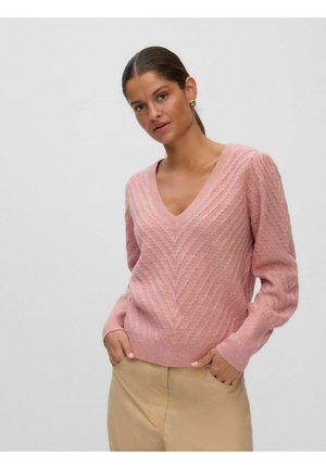 Femme portant un pull rose à col en V en maille torsadée et un pantalon beige, les mains dans les poches, sur un fond gris clair uni.