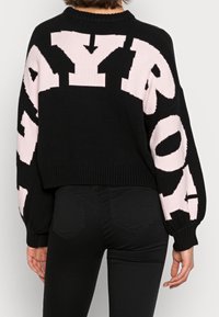 Osoba ubrana w czarny sweter z dużymi jasnoróżowymi literami na plecach i rękawach, w połączeniu z czarnymi spodniami.