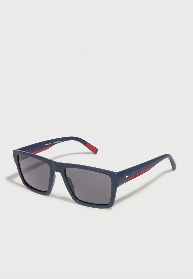 Σκούρα μπλε ορθογώνια γυαλιά ηλίου με γκρι φακούς, που διαθέτουν κόκκινες λεπτομέρειες και ένα μικρό λογότυπο της μάρκας στα πλαϊνά.