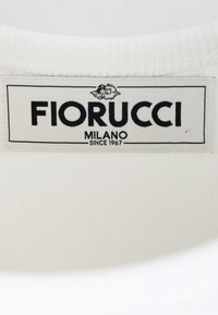 Etichetta del marchio Fiorucci su tessuto bianco, con due piccoli cherubini sopra il testo in grassetto "FIORUCCI" e "MILANO DAL 1967" sottostante.
