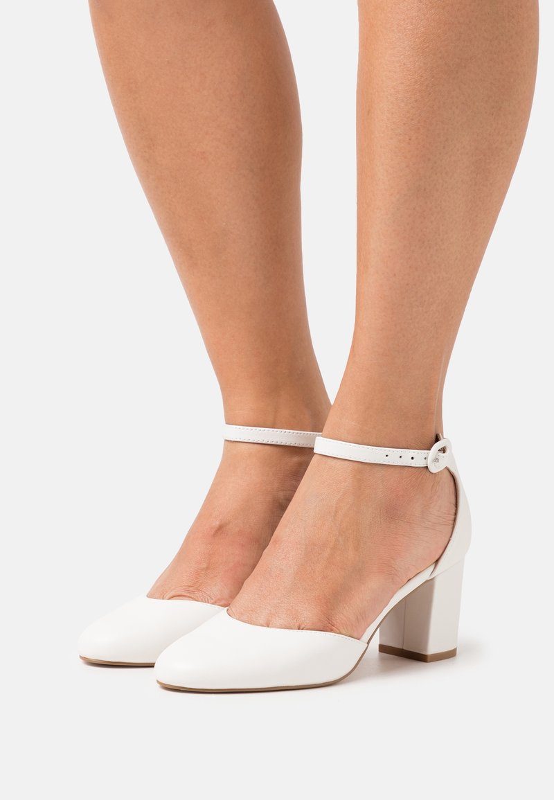 Pieds portant des talons blocs fermés blancs avec de fines brides cheville et une boucle sur un fond clair.