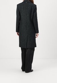 Dlouhý černý kabát z texturované látky s jemným vzorem, má jeden rozparek na zádech a propracované klopy.