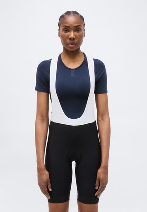 Donna con i capelli intrecciati, indossa una maglietta blu navy a maniche corte e pantaloncini da ciclismo neri con bretelle bianche, in piedi davanti a uno sfondo semplice.