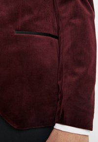 Zbliżenie na osobę noszącą bordowy aksamitny żakiet z czarnym wykończeniem kieszeni oraz białym mankietem koszuli widocznym na nadgarstku.