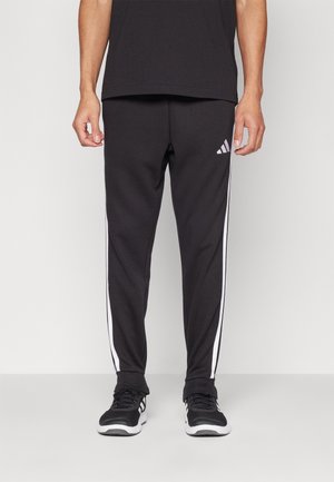 Czarne spodnie sportowe z białymi paskami po bokach, wykonane z gładkiego materiału. Posiadają logo Adidas na lewej udzie oraz elastyczny pas.