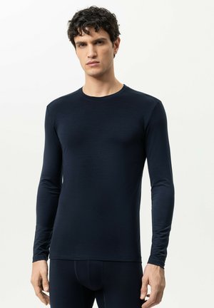Junger Mann mit lockigem dunklem Haar, trägt ein eng anliegendes schwarzes Langarmshirt und schwarze Hosen, steht vor einem schlichten hellen Hintergrund.