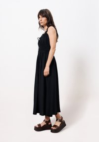 Schwarzes Maxikleid mit strukturierter Oberfläche, verstellbaren Trägern und tailliertem Oberteil, kombiniert mit braunen Keilsandalen, die eine Schnalle haben.