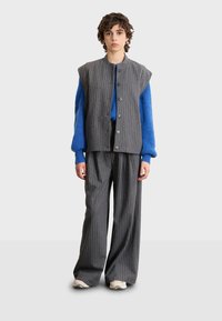 Ärmellose graue gestreifte Weste über einem blauen Stricktop, kombiniert mit lockeren grauen gestreiften Hosen und beigen Sneakers. Glatte Texturen, lässige Passform.