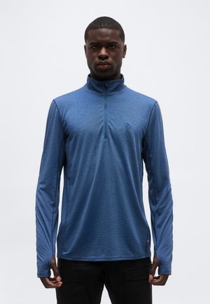 Mann trägt einen blauen langärmeligen Pullover mit halbem Reißverschluss und Daumenlöchern, steht vor einem einfarbigen weißen Hintergrund.