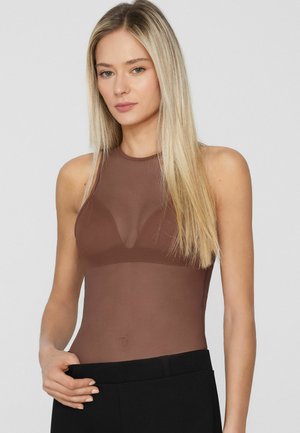 Donna bionda che indossa un top marrone trasparente senza maniche abbinato a pantaloni neri, in piedi davanti a uno sfondo grigio uniforme.