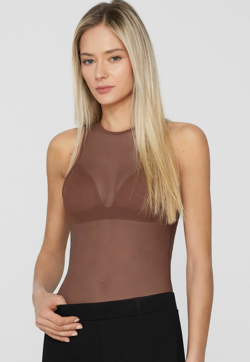 Mujer rubia con una blusa marrón translúcida sin mangas combinada con pantalones negros, posando frente a un fondo gris liso.