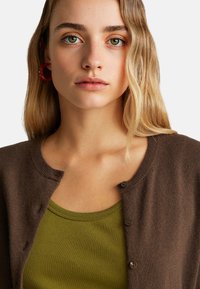 Brązowy kardigan z okrągłym dekoltem i zapięciem na guziki, założony na zielony top bez rękawów. Charakteryzuje się miękką fakturą i ciepłymi odcieniami.