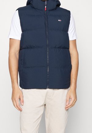 Man wearing navy blue padded vest over white short-sleeve shirt with beige pants, hands relaxed by sides, front view.