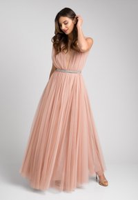 Różowa suknia z tiulu z głębokim dekoltem w kształcie litery V oraz błyszczącym srebrnym pasem w talii. Luźna, warstwowa spódnica o teksturalnym wykończeniu. Złote sandały na obcasie z paskami.