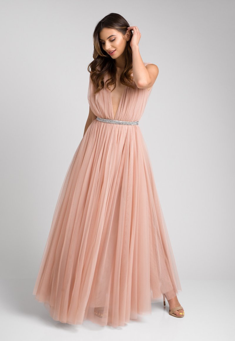 Różowa suknia z tiulu z głębokim dekoltem w kształcie litery V oraz błyszczącym srebrnym pasem w talii. Luźna, warstwowa spódnica o teksturalnym wykończeniu. Złote sandały na obcasie z paskami.