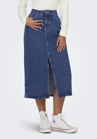 Denim midi skirt in dark blue with a front slit, high waistband, and visible stitching. Paired with white high-top sneakers.