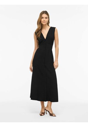 Femme portant une robe midi noire sans manches boutonnée avec un décolleté en V profond et des sandales marron à talons ouverts aux orteils, debout sur fond blanc.