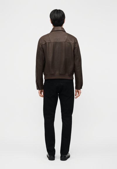 Homme portant une veste en cuir marron foncé, un pantalon noir et des chaussures noires, debout de dos sur un fond clair uni.