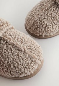 Beige tøffel med struktureret, fluffy overflade. Rund tå og blødt, tykt materiale; fleksibel gummisål for komfort og greb.