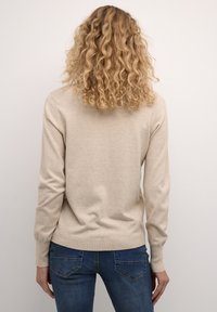 Pull en tricot beige à manches longues avec un ourlet côtelé, offrant une coupe décontractée. L'arrière présente une texture douce et un tissu agréable. Associé à un jean bleu.