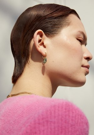 Brinco de argola dourada com um detalhe de pedra colorida, usado por uma modelo de cabelo curto. A modelo usa um suéter texturizado rosa.