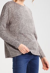 Langarmshirt aus hellbeigem Stoff mit schwarzem Sternmuster, mit lockerer Passform und abgerundetem Saum, kombiniert mit blauen Jeans.