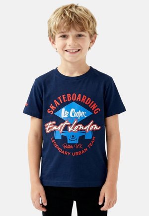 Garçon blond souriant portant un t-shirt bleu marine avec un texte et des graphiques rouges et bleus "Skateboarding East London", debout devant un fond uni.
