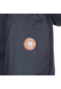 Marinblå jacka med en rund, strukturerad läderlapp där varumärkesnamnet och "sedan 1997" är präglade. Slätt tyg utan synliga mönster.