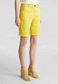 Pantalones cortos de algodón amarillos con dobladillo doblado, bolsillos laterales y cierre de botón, combinados con sandalias amarillas de tacón a juego.