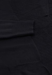 Schwarze Strickjacke mit geripptem Muster, langen Ärmeln und vorderen Taschen. Der Stoff wirkt weich und dehnbar, was den Tragekomfort erhöht.