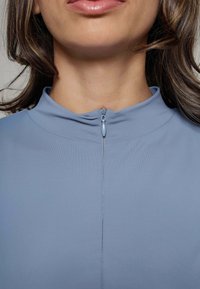 Lichtblauwe top met een hoge kraag en een ritssluiting aan de voorkant. De stof is glad met een lichte glans en heeft een eenvoudig en aansluitend ontwerp.