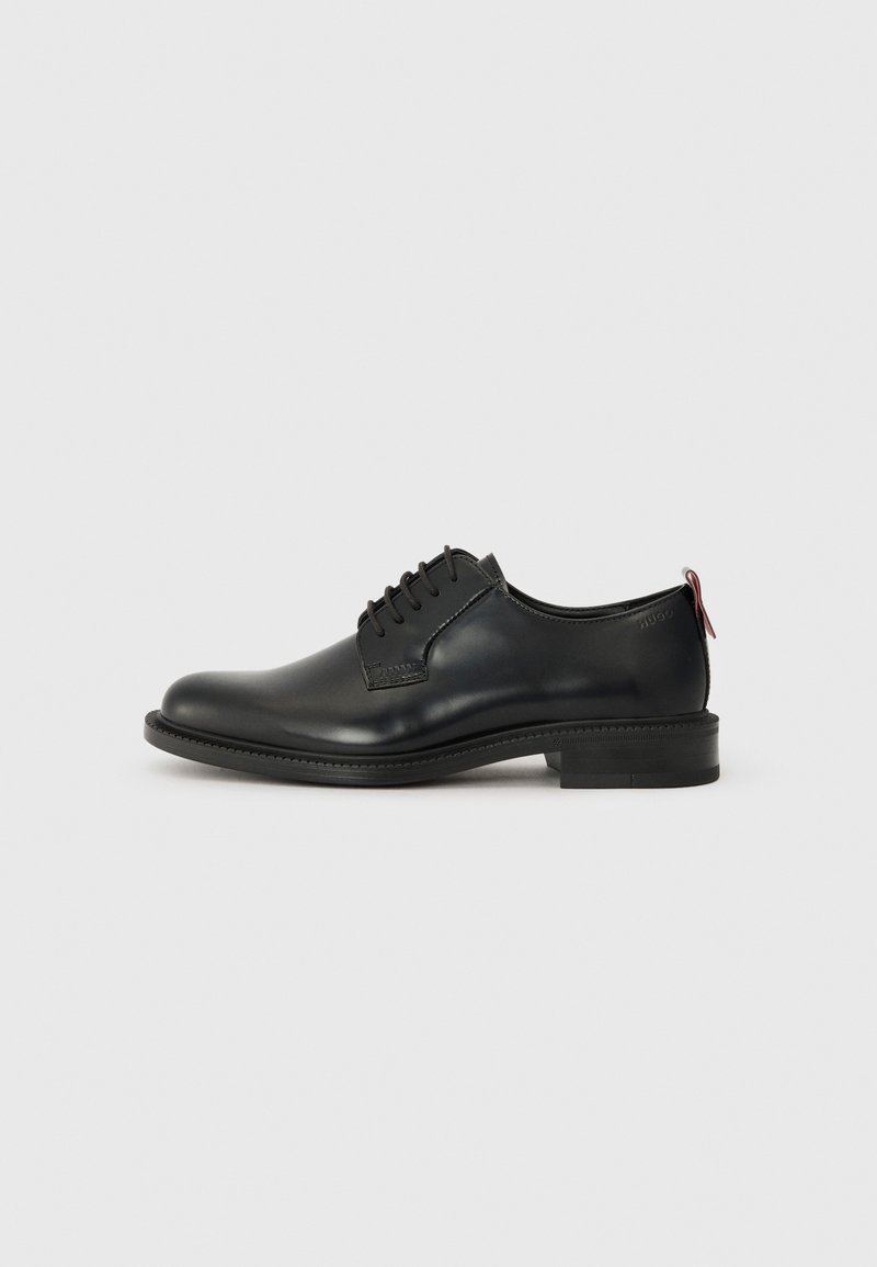 Chaussure habillée pour homme en cuir noir poli avec fermeture à lacets et petit talon, vue de profil sur fond gris.