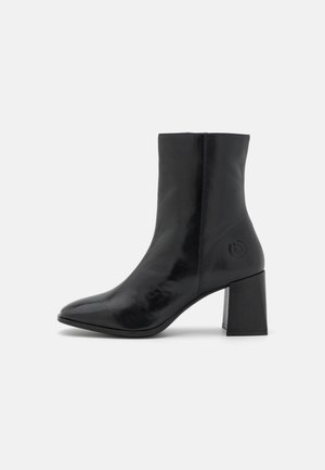 Bottine noire en cuir avec bout pointu, talon bloc et logo embossé subtil près du talon sur fond blanc uni.