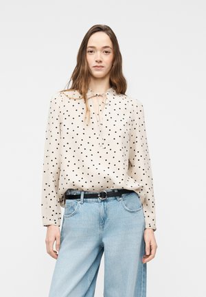 Γυναίκα με μακριά καστανά μαλλιά που φοράει εκρού μπλούζα με μαύρες βούλες και ανοιχτό μπλε ψηλόμεσο τζιν με μαύρη ζώνη.