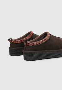 Zapatos de ante marrón sin cordones con un ribete estampado en rosa y amarillo y suela de goma negra. Bordes redondeados y acabado de superficie texturizado.