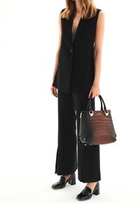 Blazer sans manches noir, pantalon large noir et chaussures noires brillantes. Tient un sac à main en croco embossé bicolore noir et marron avec des accents dorés.