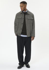 Mand står iført en ternet jakke, mørk sweater over en stribet skjorte, løse sorte bukser og sorte lædersko på hvid baggrund.