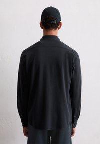 Male wearing a black long-sleeve shirt and black cap, facing away against a plain light gray textured wall background.