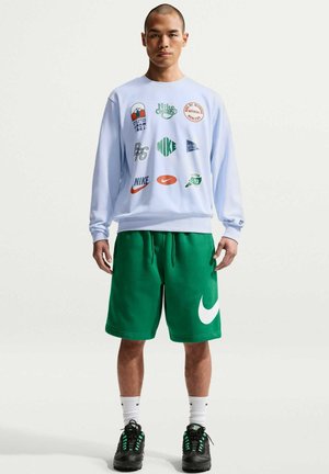 Mężczyzna stoi, ubrany w białą bluzę z kilkoma kolorowymi logo Nike, zielone szorty z dużym białym logo Nike, czarne buty, białe skarpetki.