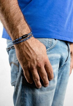 Bracciale tessuto blu e nero con chiusura nera, indossato su una mano contro un jeans azzurro chiaro con una camicia blu.