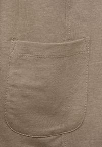 Beige kangas lenjani kwenye uso wa textured ina mfuko mkubwa uliooshwa upande mmoja, na maelezo ya seams yanayoonekana yanaboresha muundo wake.