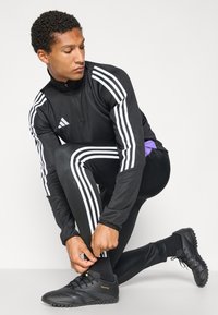 Zwart sportief trainingspak met witte strepen, met ritskraag, gecombineerd met zwarte sportschoenen. Model knielt en past de broekspijpen aan.