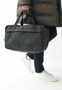 Zwarte stoffen draagtas met dubbele handvatten, een gladde textuur, een voorvak en een subtiele logopatch, vastgehouden door een persoon in een pufferjack.