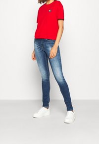 Camiseta roja corta con logo en el frontal, combinada con jeans skinny azul oscuro y zapatillas blancas con cordones. Atuendo suave, casual, de diseño simple.